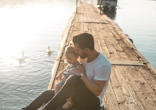 man and girl sitting on brown dock near boat and two white ducks during daytime