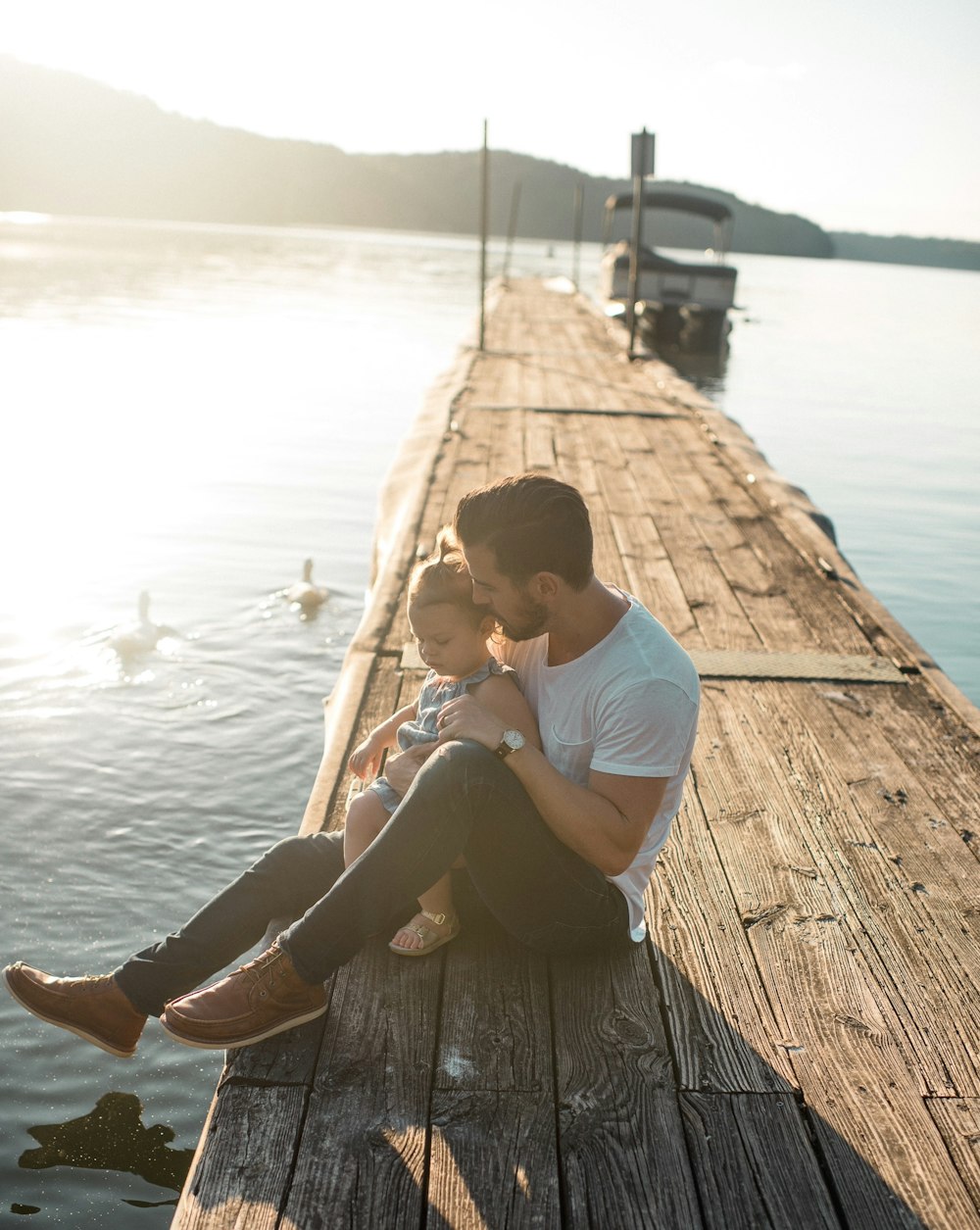 hombre y niña sentados en un muelle marrón cerca del barco y dos patos blancos durante el día