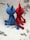 Two crocheted figures sit together, one in blue and the other in red. They have simple, rounded bodies with pointed ears or horns. Their tails are long and end with a small round tip. The background is a textured surface with scattered leaf-like patterns.