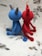 Two crocheted figures sit together, one in blue and the other in red. They have simple, rounded bodies with pointed ears or horns. Their tails are long and end with a small round tip. The background is a textured surface with scattered leaf-like patterns.
