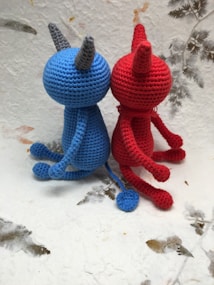 Two crocheted figures sit together, one in blue and the other in red. They have simple, rounded bodies with pointed ears or horns. Their tails are long and end with a small round tip. The background is a textured surface with scattered leaf-like patterns.