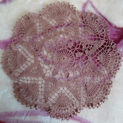A circular crochet doily with intricate patterns and floral motifs lies on a soft, fluffy surface. The delicate lacework is highlighted against the background, and the doily features a symmetrical design with scalloped edges.