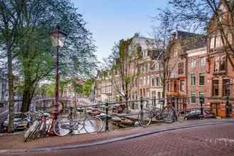A picturesque canal scene featuring traditional Dutch architecture with rows of tall, narrow houses along the water. Several bicycles are parked on a bridge, which is adorned with classic street lamps. Leafy trees line the canal, adding greenery to the scene.