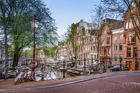 A picturesque canal scene featuring traditional Dutch architecture with rows of tall, narrow houses along the water. Several bicycles are parked on a bridge, which is adorned with classic street lamps. Leafy trees line the canal, adding greenery to the scene.