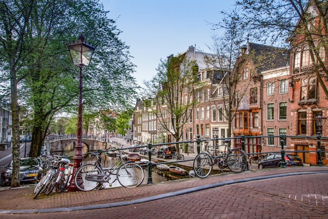 A picturesque canal scene featuring traditional Dutch architecture with rows of tall, narrow houses along the water. Several bicycles are parked on a bridge, which is adorned with classic street lamps. Leafy trees line the canal, adding greenery to the scene.