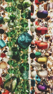 A collection of colorful beads strung together in various shapes and sizes. The beads display a multitude of colors, predominantly greens, reds, and purples, with a variety of textures ranging from smooth to faceted.