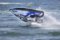 A person performing a stunt with a jet ski on water, partially submerged as the vehicle flips over while creating a splash.