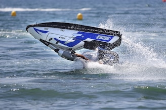 A person performing a stunt with a jet ski on water, partially submerged as the vehicle flips over while creating a splash.