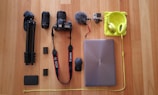 A collection of electronic and photography gear neatly arranged on a wooden floor, including a tripod, two camera lenses, a Canon camera, a microphone, a GoPro, a laptop, a mouse, headphones, and various accessories.