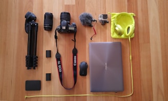 A collection of electronic and photography gear neatly arranged on a wooden floor, including a tripod, two camera lenses, a Canon camera, a microphone, a GoPro, a laptop, a mouse, headphones, and various accessories.