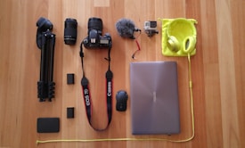 A collection of electronic and photography gear neatly arranged on a wooden floor, including a tripod, two camera lenses, a Canon camera, a microphone, a GoPro, a laptop, a mouse, headphones, and various accessories.
