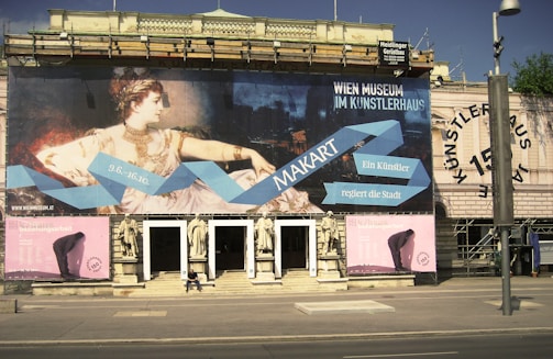 A large museum banner is displayed on the exterior of a historical building. The banner features a classical artwork of a woman in elegant attire and includes text promoting a museum event, with a blue geometric design leading the eye across the display. Two smaller pink posters flank the entrance and feature abstract art. Several statues are placed above the entrance, and a person is seated on the steps, providing a sense of scale.