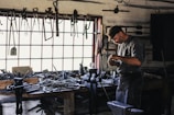 A craftsman or blacksmith is working in a rustic workshop filled with tools hanging from the wall and cluttered on a wooden workbench. He is focused on his work, wearing a cap and apron, and surrounded by iron tools and equipment.