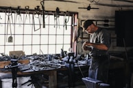 A craftsman or blacksmith is working in a rustic workshop filled with tools hanging from the wall and cluttered on a wooden workbench. He is focused on his work, wearing a cap and apron, and surrounded by iron tools and equipment.