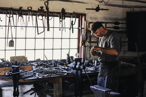 A craftsman or blacksmith is working in a rustic workshop filled with tools hanging from the wall and cluttered on a wooden workbench. He is focused on his work, wearing a cap and apron, and surrounded by iron tools and equipment.
