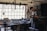 A craftsman or blacksmith is working in a rustic workshop filled with tools hanging from the wall and cluttered on a wooden workbench. He is focused on his work, wearing a cap and apron, and surrounded by iron tools and equipment.