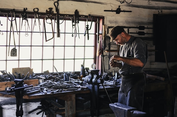 A craftsman or blacksmith is working in a rustic workshop filled with tools hanging from the wall and cluttered on a wooden workbench. He is focused on his work, wearing a cap and apron, and surrounded by iron tools and equipment.