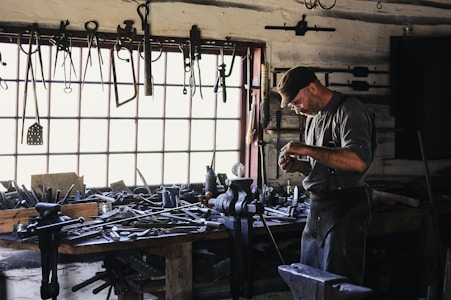 A craftsman or blacksmith is working in a rustic workshop filled with tools hanging from the wall and cluttered on a wooden workbench. He is focused on his work, wearing a cap and apron, and surrounded by iron tools and equipment.