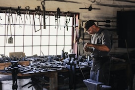 A craftsman or blacksmith is working in a rustic workshop filled with tools hanging from the wall and cluttered on a wooden workbench. He is focused on his work, wearing a cap and apron, and surrounded by iron tools and equipment.