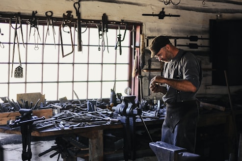 A craftsman or blacksmith is working in a rustic workshop filled with tools hanging from the wall and cluttered on a wooden workbench. He is focused on his work, wearing a cap and apron, and surrounded by iron tools and equipment.