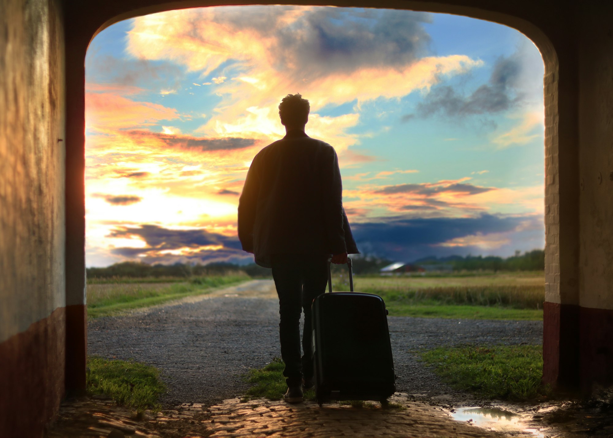 homme sortant d'un tunnel à contre-jour et tenant une valise devant un levé de soleil avec des nuages
