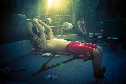 A shirtless man in red shorts lies on a bench press, lifting a barbell with the help of a spotter. Another person is sitting in the background, resting on a bench. Sunlight filters through the trees, casting a moody and serene atmosphere in the outdoor gym setting.