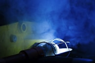 A light source enclosed in a protective metal cage emits a glow, casting shadows on a surface. Blue smoke or mist fills the air, adding a mysterious and dramatic effect. A bolt and circular opening are visible on the textured yellow surface in the background.
