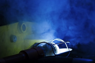 A light source enclosed in a protective metal cage emits a glow, casting shadows on a surface. Blue smoke or mist fills the air, adding a mysterious and dramatic effect. A bolt and circular opening are visible on the textured yellow surface in the background.