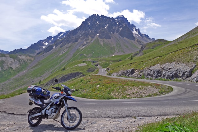 A winding mountain road stretching into the distance with a single motorcycle parked at a scenic overlook under a bright blue sky.