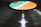 A close-up view of a vinyl record on a record player, with the label area colored in shades of turquoise, white, and orange. The grooves of the record reflect light, creating a shiny, smooth surface.
