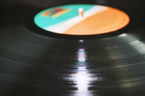 A close-up view of a vinyl record on a record player, with the label area colored in shades of turquoise, white, and orange. The grooves of the record reflect light, creating a shiny, smooth surface.