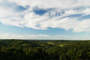A scenic landscape photograph featuring a vast view of rolling hills covered in lush green forests under a sky with dramatic, white clouds scattered across a bright blue background. The horizon extends far into the distance, capturing a serene and expansive natural setting.