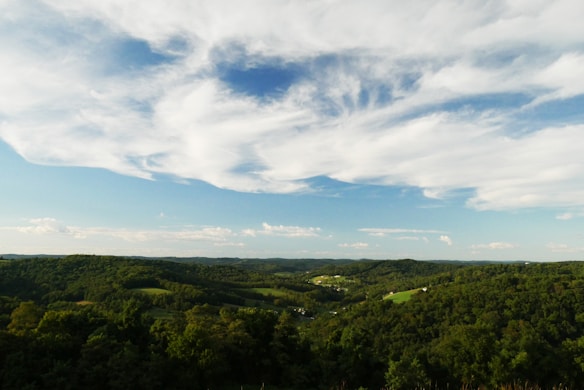 A scenic landscape photograph featuring a vast view of rolling hills covered in lush green forests under a sky with dramatic, white clouds scattered across a bright blue background. The horizon extends far into the distance, capturing a serene and expansive natural setting.
