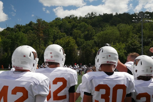 A group of American football players wearing white jerseys and helmets stands on a field, facing away from the camera. The background shows a lush, green forest under a partly cloudy blue sky.