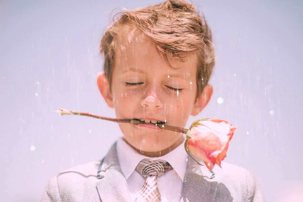 A little boy in a formal suit holding a pink rose between his teeth