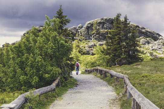 A scenic pathway winds through lush green vegetation, bordered by wooden fences, leading towards rocky hills under a cloudy sky. A person with a backpack is walking along the path, emphasizing the tranquility and natural beauty of the setting.