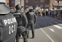 Several police officers stand in the foreground, dressed in black uniforms with helmets and tactical gear. They face a crowd gathered in the distance, blurred in the background. The scene is set on a city street with brick buildings nearby and traffic signs visible.