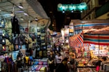 A busy street market at night, with numerous stalls covered by striped tarps, displaying an array of items such as bags, backpacks, and colorful suitcases. A diverse crowd of people navigates through the market, exploring different stalls under the bright lights. Signs with Chinese characters can be seen above the stalls.