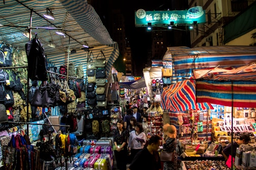 A busy street market at night, with numerous stalls covered by striped tarps, displaying an array of items such as bags, backpacks, and colorful suitcases. A diverse crowd of people navigates through the market, exploring different stalls under the bright lights. Signs with Chinese characters can be seen above the stalls.