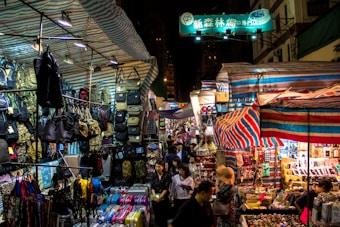 A busy street market at night, with numerous stalls covered by striped tarps, displaying an array of items such as bags, backpacks, and colorful suitcases. A diverse crowd of people navigates through the market, exploring different stalls under the bright lights. Signs with Chinese characters can be seen above the stalls.