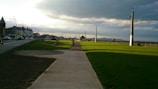 A pathway stretches ahead along a coastal promenade with green grass on one side and a road lined with buildings on the other. Streetlights and poles stand at intervals, and the sky is partially cloudy, casting shadows and sunlight across the scene. In the distance, a small building and distant hills are visible.