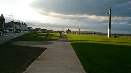 A pathway stretches ahead along a coastal promenade with green grass on one side and a road lined with buildings on the other. Streetlights and poles stand at intervals, and the sky is partially cloudy, casting shadows and sunlight across the scene. In the distance, a small building and distant hills are visible.