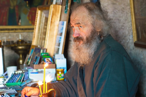 An elderly man with a long, gray beard is seated in a dimly lit room surrounded by various objects, including books, papers, and lit candles. The man has a distant expression and is wearing a dark coat. The background is filled with shelves, religious icons, and frames.