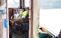 An old, weathered helm of a boat with a wooden steering wheel, surrounded by various objects including a yellow container, a red lever, and some nautical instruments. The cabin is rustic with visible wear and tear, suggesting frequent use. The scene outside the cabin shows an overcast sky and an expanse of water.