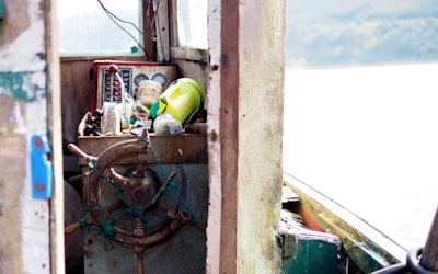 An old, weathered helm of a boat with a wooden steering wheel, surrounded by various objects including a yellow container, a red lever, and some nautical instruments. The cabin is rustic with visible wear and tear, suggesting frequent use. The scene outside the cabin shows an overcast sky and an expanse of water.
