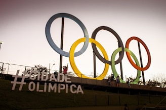 Brightly colored, large Olympic rings are mounted on an outdoor structure against a dusky sky. People are visible interacting with the installation, capturing the essence of a festive, sports-oriented environment. The hashtag sign '#Cidade Olimpica' is prominently displayed in the foreground, suggesting the location's link to Olympic activities.