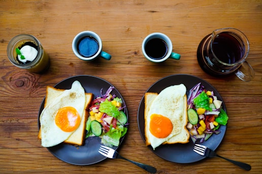 A wooden table displays a neatly arranged breakfast setup for two. Each setting has a plate with a slice of toast topped with a sunny-side-up egg, accompanied by a colorful salad featuring lettuce, cucumber, red cabbage, and diced fruit. There are two mugs of black coffee and a glass pitcher filled with coffee as well. Additionally, a jar containing a layered dessert with a dollop of cream is present.