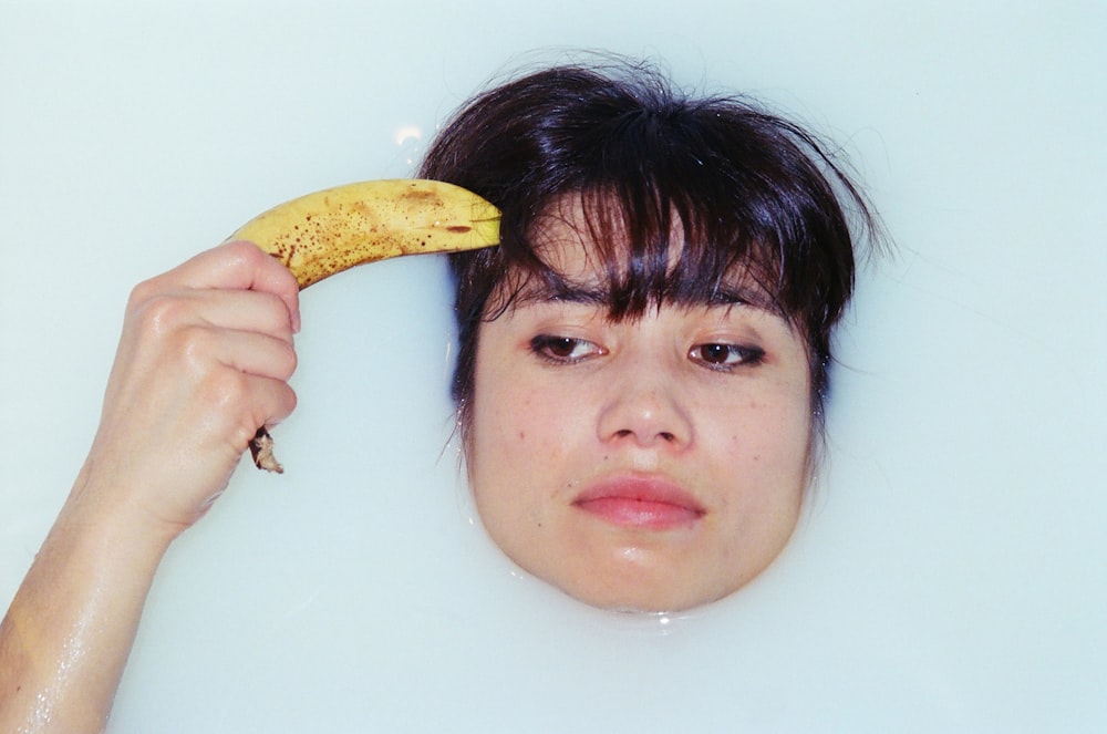 Una mujer sosteniendo un plátano en su cabeza mientras está completamente sumergida en una bañera.