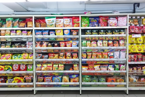 A supermarket aisle filled with a variety of colorful packaged snacks arranged on metal shelving units. Different brands and flavors of chips and other snacks are prominently displayed, creating a vibrant and diverse display of products.