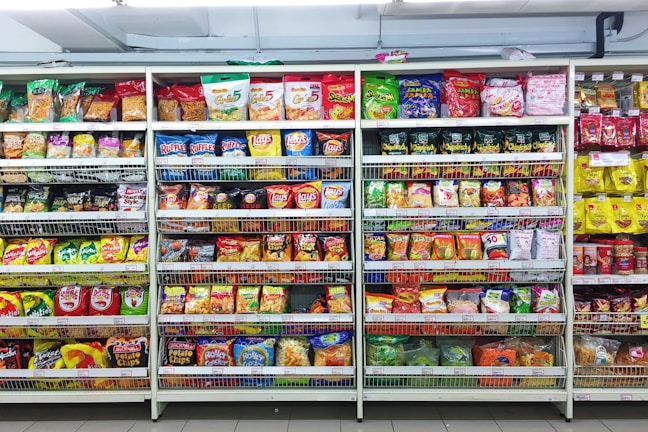 A supermarket aisle filled with a variety of colorful packaged snacks arranged on metal shelving units. Different brands and flavors of chips and other snacks are prominently displayed, creating a vibrant and diverse display of products.